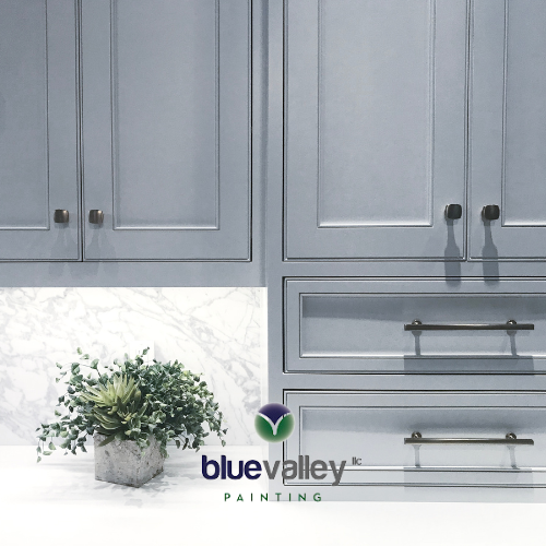 Close-up of freshly painted kitchen cabinets in a soft gray color with brushed brass hardware. A small potted plant sits on the countertop with a white countertop and backsplash.