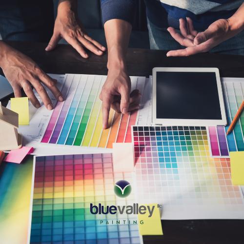 A group of people are gathered around a table, discussing color palettes and design ideas. Paint chips, a tablet, and a house model are spread out on the table.